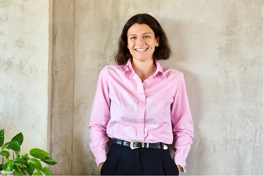 A smiling woman with shoulder-length dark hair wearing a light pink button-up shirt and dark trousers.